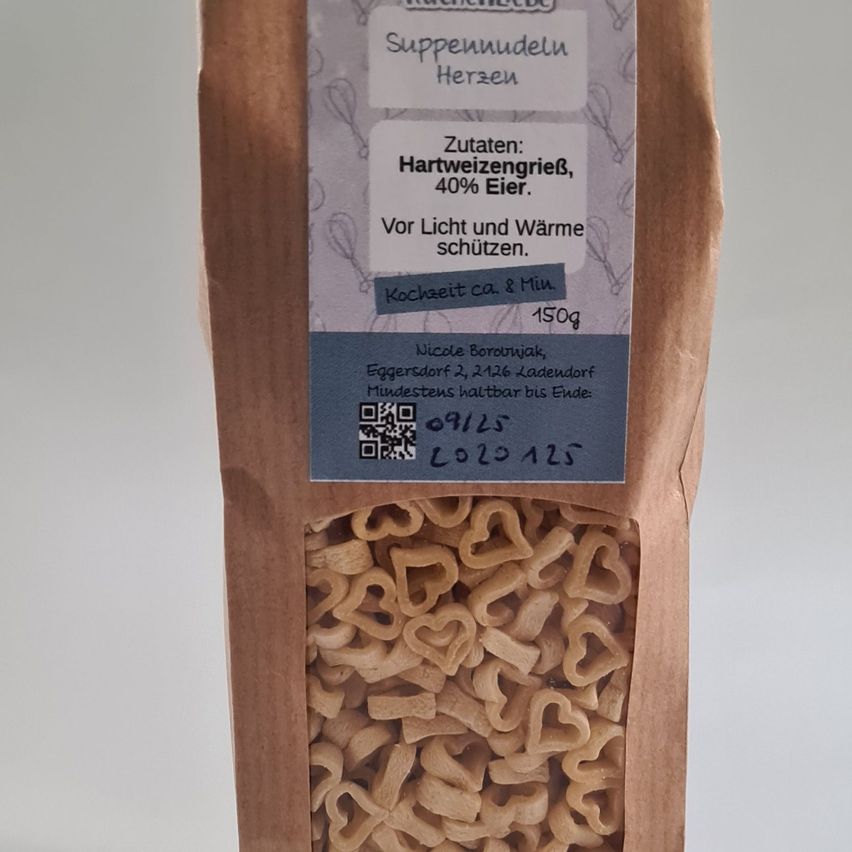 Ein brauner Beutel mit einem Etikett, das 'Suppennudeln Herzen' und herzförmige Nudeln im Inneren anzeigt. Das Etikett erwähnt auch Zutaten wie Weizenmehl und Ei sowie die Kochzeit. Der Beutel ist vor Licht und Wärme geschützt.
