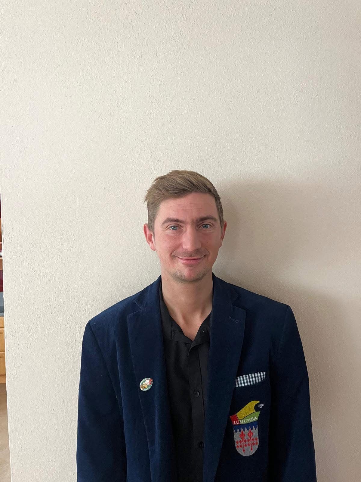 Ein Mann mit einem Abzeichen auf seinem Blazer steht gegen eine weiße Wand, lächelt und schaut in die Kamera.