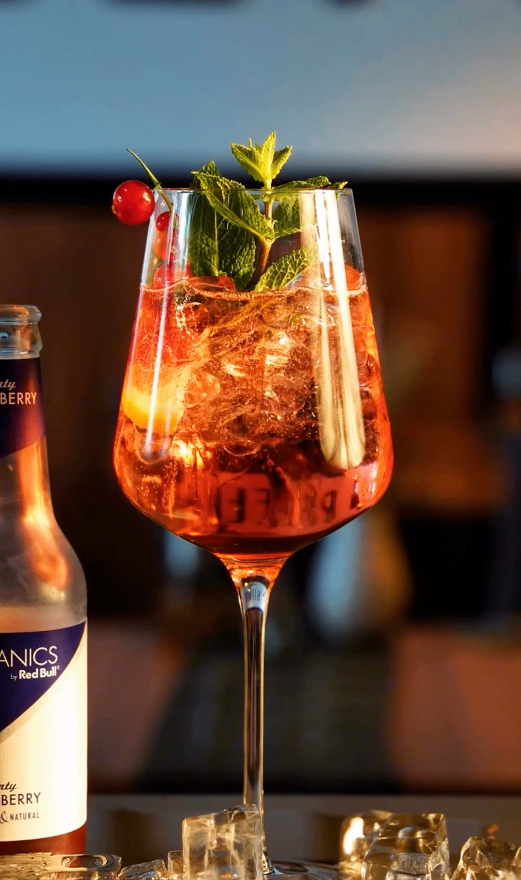 Ein Nahaufnahme eines Cocktailglases mit rotem Inhalt, mit Minzblättern und einer Kirsche garniert. Daneben steht eine Flasche Red Bull Energy Drink.