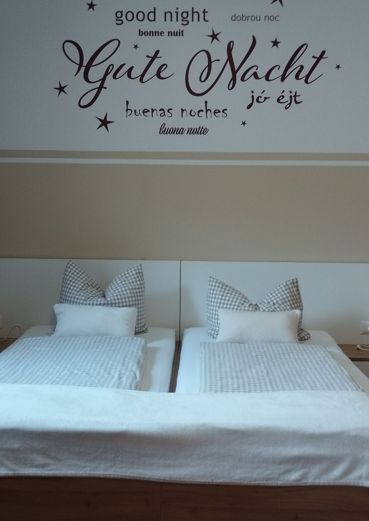Zwei Einzelbetten in einem Zimmer mit einer weißen Wand und einem dekorativen Wandaufkleber, der 'Gute Nacht' in verschiedenen Sprachen sagt.