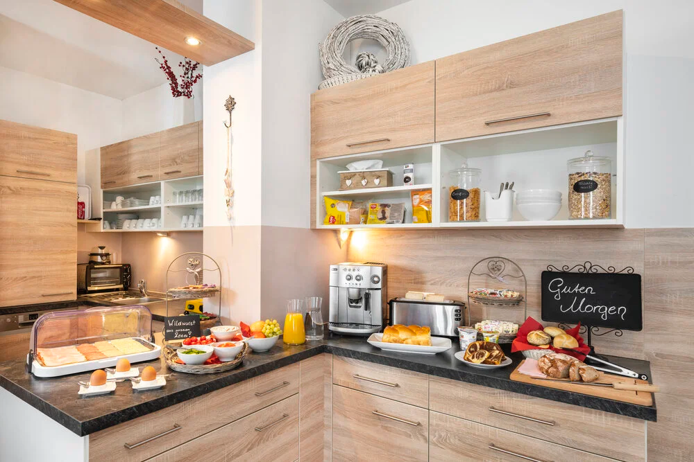A well-equipped kitchen with a breakfast buffet on the countertop. A coffee machine and toaster are visible, along with various food items. The cabinets and shelves hold plates, bowls, and snacks. A decorative wreath and plant add a touch of nature.