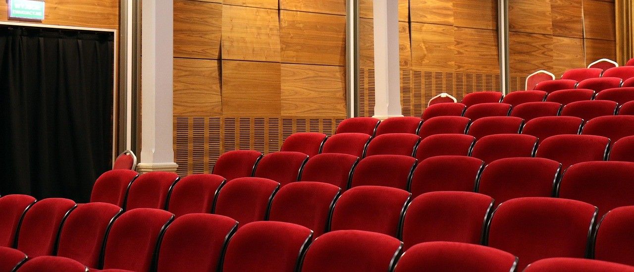 Ein leerer Saal mit Holzwänden und roten Stühlen. Die Stühle sind in Reihen angeordnet, und es gibt zwei weiße Säulen in der Mitte.