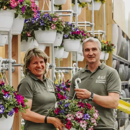 Zwei Personen in grünen Uniformen, möglicherweise Angestellte, stehen neben einer Wand mit hängenden Topfpflanzen. Sie lächeln und posieren für ein Foto.
