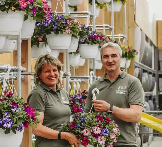 Zwei Personen in grünen Uniformen, möglicherweise Angestellte, stehen neben einer Wand mit hängenden Topfpflanzen. Sie lächeln und posieren für ein Foto.