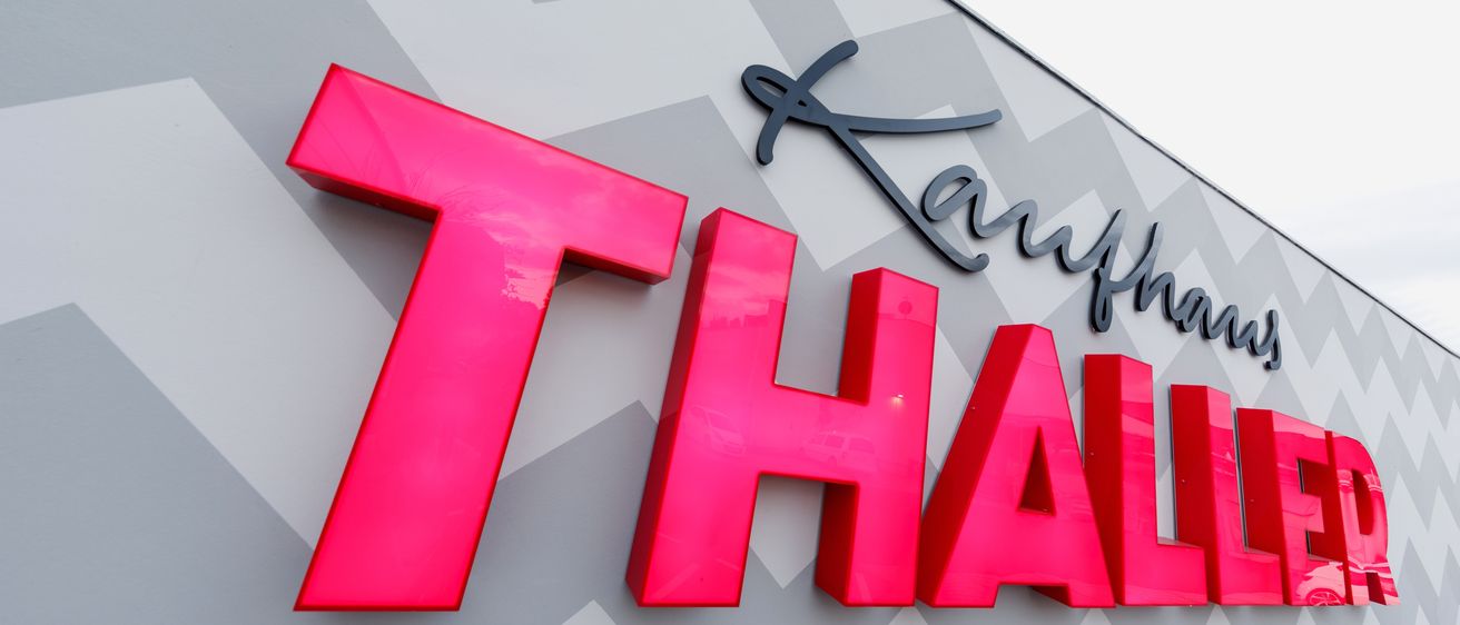 Ein leuchtend pinkes Schild mit dem Wort 'HALL' auf einer grauen chevron-gemusterten Wand. Über den Buchstaben steht 'Karnhaus' in schwarz geschrieben.