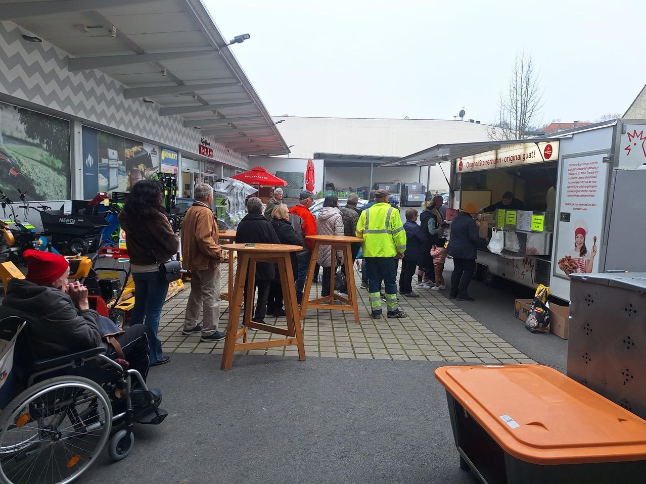 Eine Gruppe von Menschen steht und unterhält sich in der Nähe von Tischen und einem Essenswagen. Einige tragen Jacken und Mützen. Rechts befindet sich ein Aufbewahrungsbehälter.