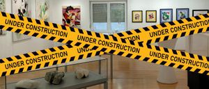 Eine Kunstgalerie mit einem Baustellenband über dem Eingang. Kunstwerke hängen an der Wand, und ein Glastisch mit Felsen steht im Vordergrund.