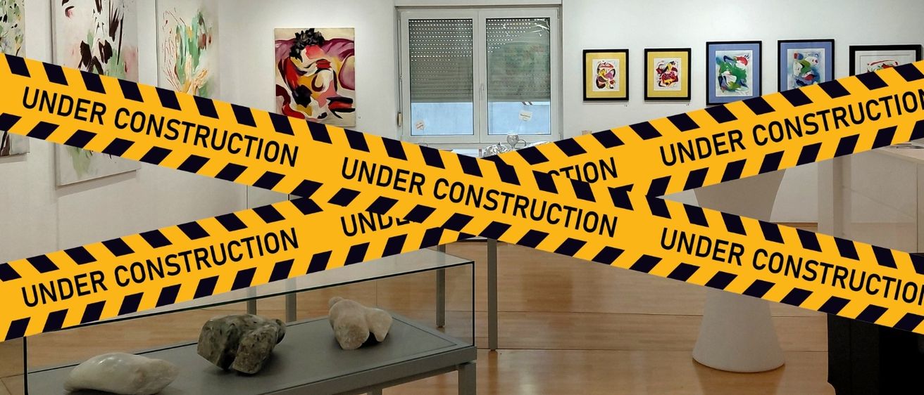 Eine Kunstgalerie mit einem Baustellenband über dem Eingang. Kunstwerke hängen an der Wand, und ein Glastisch mit Felsen steht im Vordergrund.