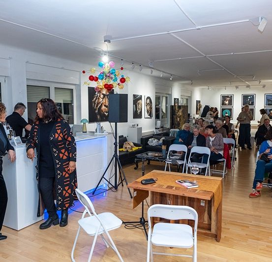 Eine Gruppe von Menschen versammelt sich in einer Kunstgalerie, stehend und sitzend auf Stühlen. Eine Frau spricht in ein Mikrofon an einem Schreibtisch, während eine andere Frau in der Nähe steht. Kunstwerke hängen an den Wänden.