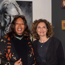 Zwei Frauen stehen in einer Kunstgalerie, lächeln und posieren für ein Foto vor einem großen schwarz-weißen Porträt.