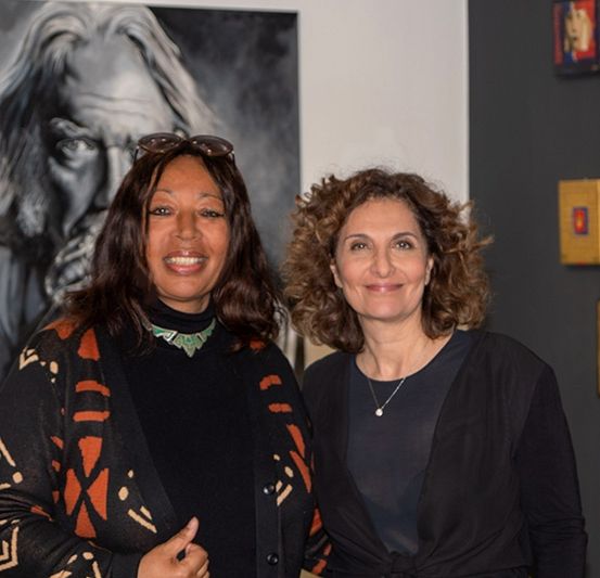 Zwei Frauen stehen in einer Kunstgalerie, lächeln und posieren für ein Foto vor einem großen schwarz-weißen Porträt.