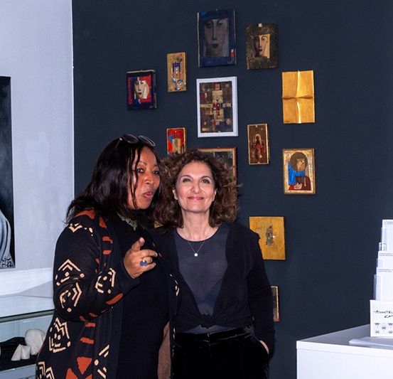 Zwei Frauen stehen nebeneinander vor einer Wand mit verschiedenen Gemälden und Skulpturen. Eine Frau zeigt auf etwas vor sich, während die andere lächelt.