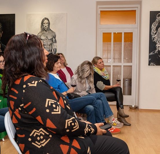 Eine Gruppe von Frauen versammelt sich in einer Kunstgalerie, schaut sich eine Präsentation an und sitzt auf Stühlen. Gemälde schmücken die Wände.