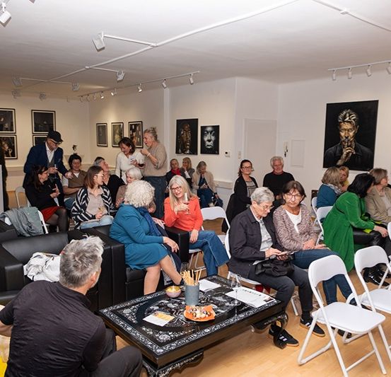 Eine Gruppe von Menschen sitzt in Stühlen in einem Raum mit weißen Wänden, umgeben von Gemälden. Der Raum hat weiße Deckenleuchten und einen schwarzen Kaffeetisch. Die Leute schauen auf einen Mann auf der linken Seite.