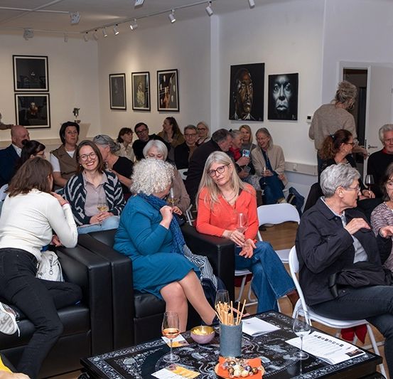 Ein Raum voller Erwachsener, die in Stühlen und Sofas sitzen und sich unterhalten. Ein Tisch mit Weingläsern und Essen steht in der Mitte. Kunstrahmen hängen an den Wänden.