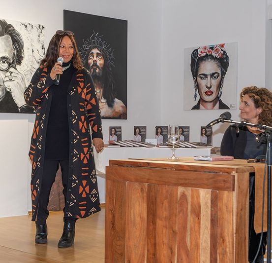 Eine Frau steht in einer Kunstgalerie mit einem Mikrofon und spricht. Hinter ihr hängen Kunstwerke an den Wänden. Eine andere Frau sitzt an einem Schreibtisch mit einem Buch und einem Weinglas.