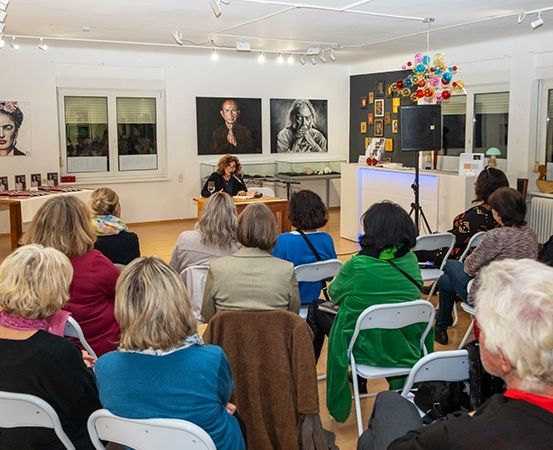 Eine Gruppe von Menschen sitzt in einer Kunstgalerie. Eine Frau steht vor einem Schreibtisch mit einem Weinglas. Mehrere Gemälde hängen an der Wand.