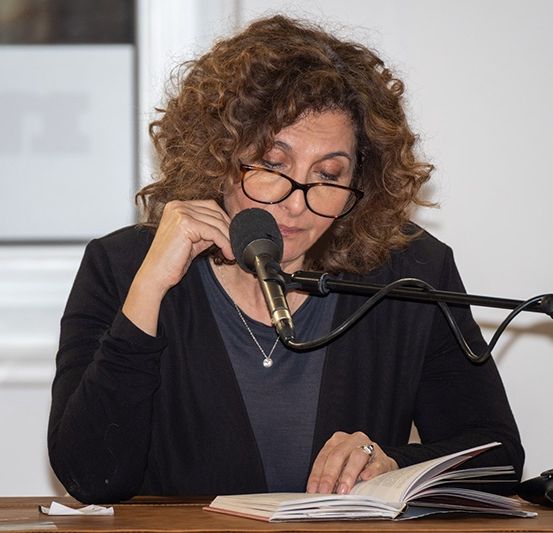 Eine Frau liest aus einem Buch in ein Mikrofon. Sie trägt eine Brille und eine schwarze Jacke. Ein Glas Wasser steht auf dem Tisch.