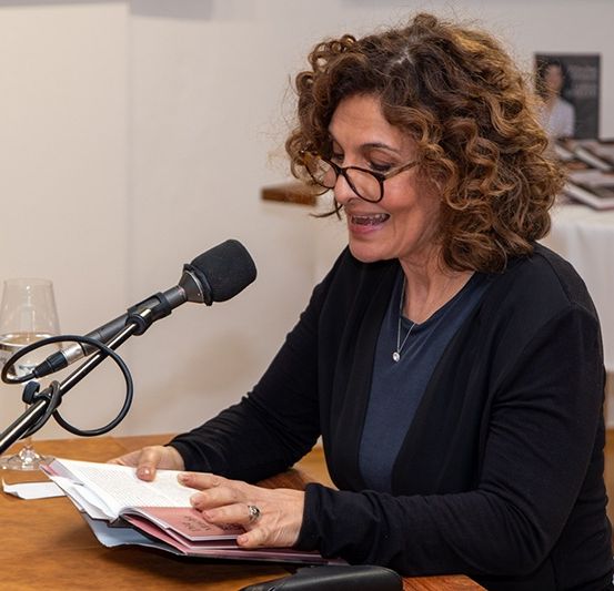 Eine Frau mit Brille und Ring liest von einem Buch, während sie an einem Tisch sitzt, der ein Mikrofon und ein Weinglas aufweist. Dahinter zeigt ein Tisch mehrere Bücher.