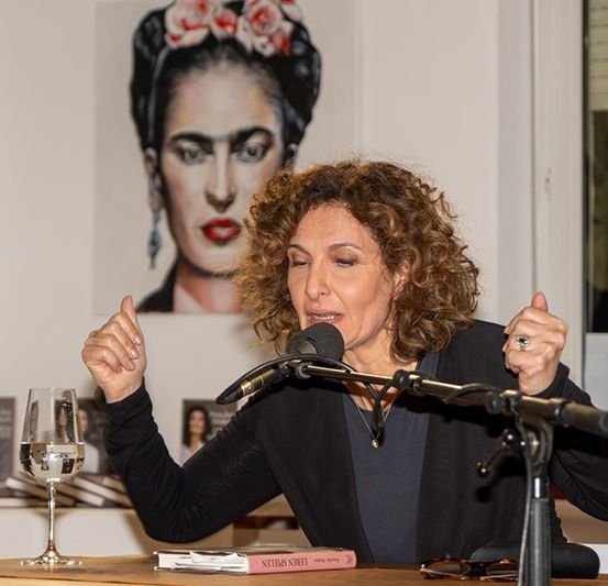 Eine Frau spricht in ein Mikrofon und gestikuliert mit ihren Händen. Hinter ihr ist ein Gemälde einer Frau mit roten Lippen und Blumen im Haar.