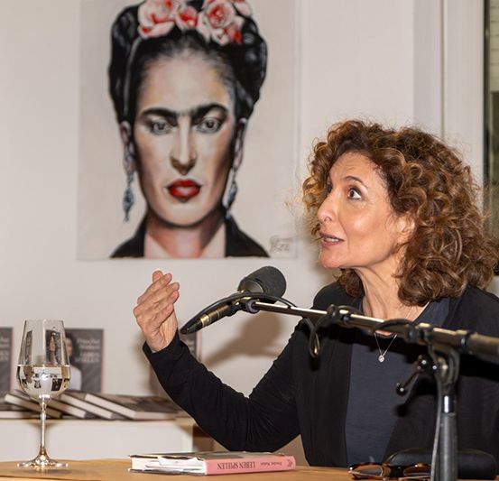 Eine Frau spricht an einem Podium mit einem Mikrofon, das ein schwarzes Oberteil trägt. Vor ihr steht ein Weinglas auf einem Tisch. Hinter ihr befindet sich ein Poster von Frida Kahlo.