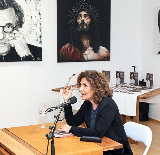 Eine Frau sitzt an einem Holzschreibtisch mit einem Buch und einem Mikrofon. Hinter ihr hängen drei große Gemälde an der Wand. Eines stellt Jesus dar, ein anderes einen Mann mit Brille und das letzte eine Frau mit Blumen.