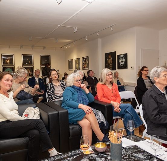 Eine Gruppe von Menschen sitzt in einem Kunstgalerieraum. Einige tragen Brillen, andere haben Taschen. Der Raum hat Kunstwerke an der Wand. Ein Tisch hat Weingläser und eine Schüssel. Eine Frau hält eine Kamera.