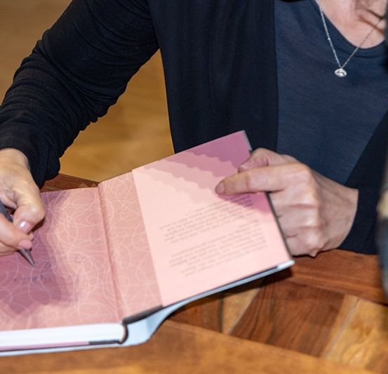 Eine Person signiert ein Buch mit einem Stift auf einem Holztisch. Ein Mikrofon steht vor ihr.