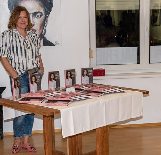Eine Frau steht bei einem Tisch mit Büchern, ein wandmontiertes Porträt hinter ihr. Der Raum hat Holzböden und Fenster mit Jalousien.