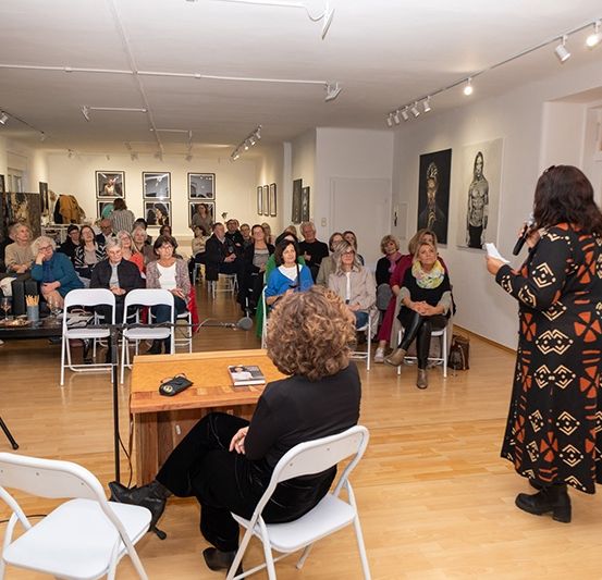 Ein Ausstellungsraum mit einem vielfältigen Publikum, das auf Stühlen sitzt, eine Frau spricht in ein Mikrofon an einem Schreibtisch. Kunstwerke hängen an den Wänden und ein Lautsprecher steht links.