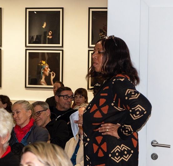 Eine Frau steht vor einer Gruppe in einer Kunstgalerie und hält möglicherweise eine Rede. Das Publikum hört aufmerksam zu, mit gerahmten Gemälden an der Wand im Hintergrund.