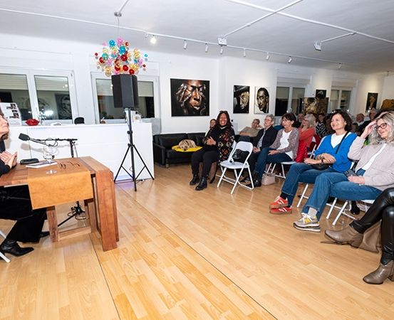 Eine Frau spricht an einem Rednerpult in einem Raum voller Stühle. Das Publikum sitzt auf dem Boden, und ein Lautsprecher und ein Mikrofon sind aufgebaut. Kunstwerke schmücken die Wände.