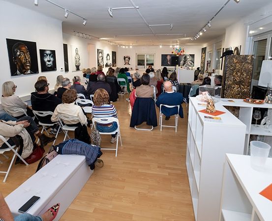 Ein Raum voller Kunstwerke an den Wänden und einer Menschenmenge in Stühlen. Die Menschen versammeln sich um eine Bühne mit einem Podium. Einige Gegenstände stehen auf dem Tisch und eine Lampe ist an der Decke montiert. Ein Mülleimer steht rechts.