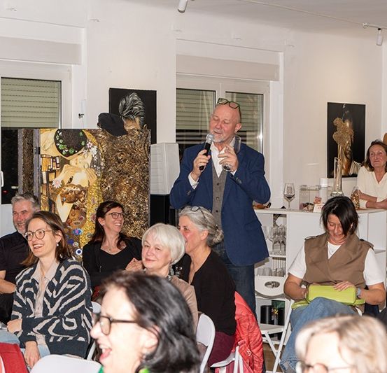 Ein Mann spricht in ein Mikrofon in einem Raum mit mehreren sitzenden und stehenden Personen, von denen einige lächeln, mit Gemälden an der Wand.