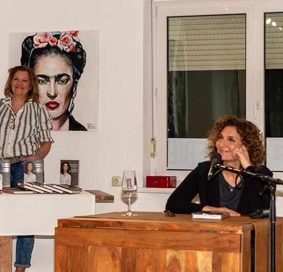 Zwei Frauen an einem Podium, eine sitzt mit einem Buch, die andere steht. Ein Gemälde von Frida Kahlo und Bücher sind im Hintergrund. Die Szene spielt sich in einem Raum mit Fenstern und Jalousien ab.
