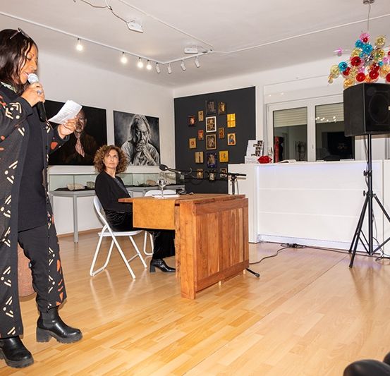 Eine Frau spricht in ein Mikrofon in einem Raum mit Holzböden, umgeben von Kunstwerken, während eine andere Frau zuhört.