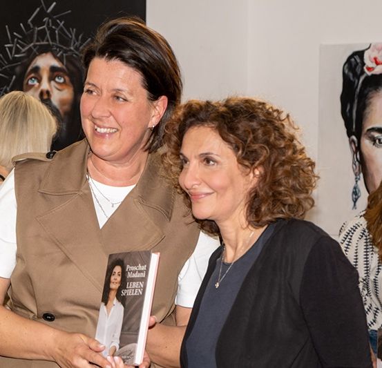 Zwei Frauen lächeln und halten ein Buch in einer Galerie. Dahinter schaut eine weitere Frau auf ein Buch. Links steht ein Mann und schaut auf das Buch. An der Wand befinden sich Gemälde von Jesus Christus und einer Frau.