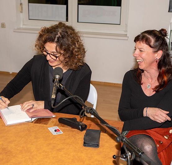 Zwei Frauen sitzen an einem Tisch mit einem Mikrofon, eine schreibt in ein Buch und die andere lächelt. Ein Weinglas und ein Notizbuch liegen auf dem Tisch. Hinter ihnen sind Fenster und ein Schaukasten mit einem Stein darin zu sehen.