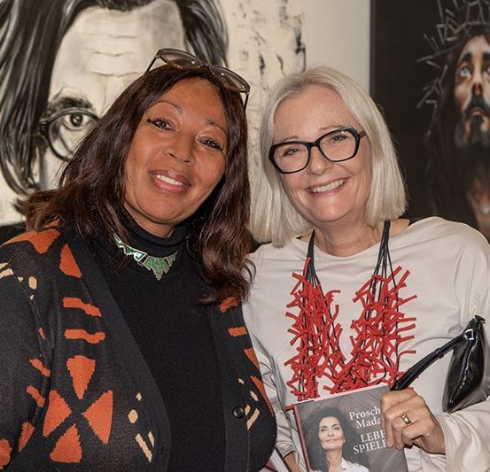 Zwei Frauen stehen lächelnd eng beieinander vor einer Kunstgalerie mit Porträts. Eine Frau hält ein Buch.