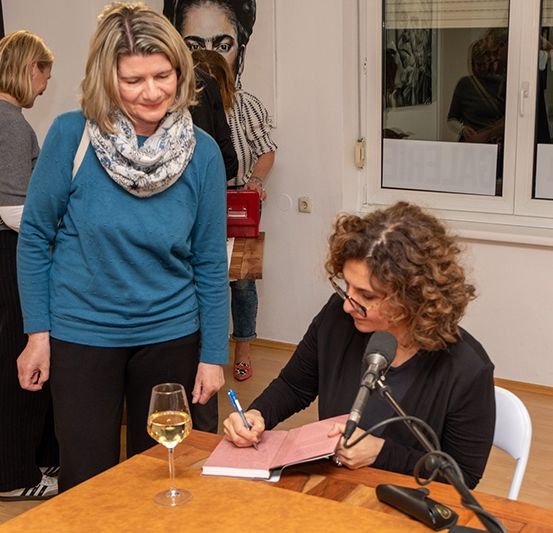 Eine Frau signiert ein Buch bei einer Veranstaltung. Sie sitzt an einem Tisch mit Mikrofon und einem Glas Wein. Eine andere Frau steht hinter ihr und lächelt.