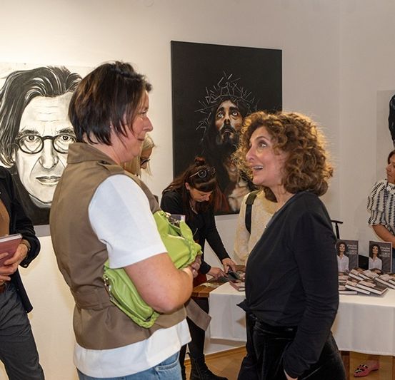 Leute in einer Kunstgalerie diskutieren, während zwei Frauen Bücher auf einem Tisch betrachten. Ein Gemälde zeigt Jesus.