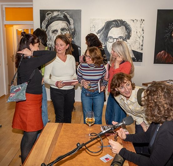 Eine Gruppe von Frauen versammelt sich um einen Tisch, eine von ihnen hält ein Mikrofon, vielleicht in einer Kunstgalerie.