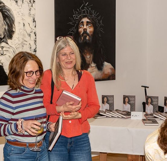 Zwei Frauen stehen in einer Kunstgalerie, eine hält ein Buch, die andere einen Weinglas. Gemälde von Jesus und Frida Kahlo an der Wand.