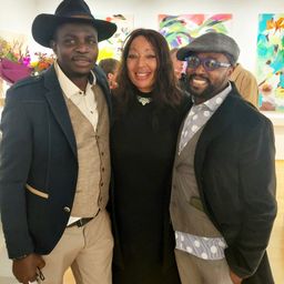 Drei Menschen posieren für ein Foto in einer Kunstgalerie. Zwei Männer und eine Frau stehen eng zusammen und lächeln. Der Mann auf der linken Seite trägt einen Hut und eine Weste, während der Mann auf der rechten Seite eine Brille und einen Mantel trägt.