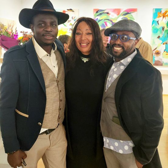 Drei Menschen posieren für ein Foto in einer Kunstgalerie. Zwei Männer und eine Frau stehen eng zusammen und lächeln. Der Mann auf der linken Seite trägt einen Hut und eine Weste, während der Mann auf der rechten Seite eine Brille und einen Mantel trägt.