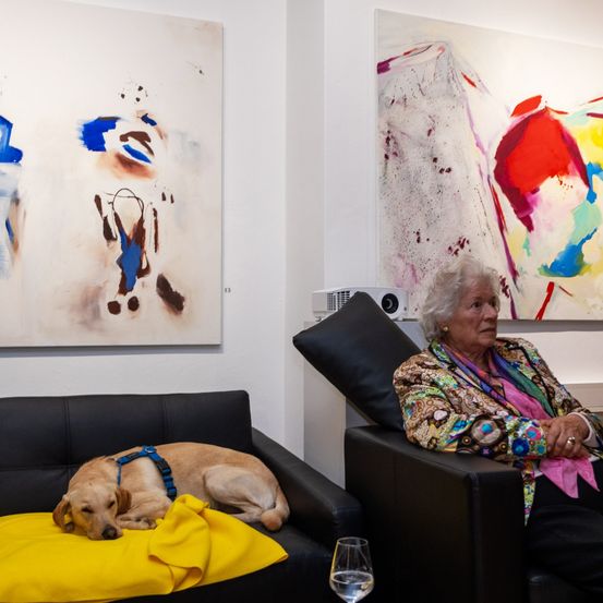 Eine ältere Frau sitzt in einem Sessel mit einem schlafenden Hund auf einem gelben Kissen. Hinter ihnen hängen zwei große abstrakte Gemälde an der Wand.