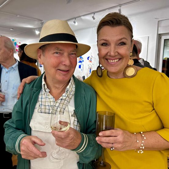 Ein Mann und eine Frau bei einer Veranstaltung halten Weingläser und lächeln. Der Mann trägt einen Hut und eine Schürze. Hinter ihnen steht ein Mann in einem Anzug in der Nähe einer Wand mit Kunstwerken.