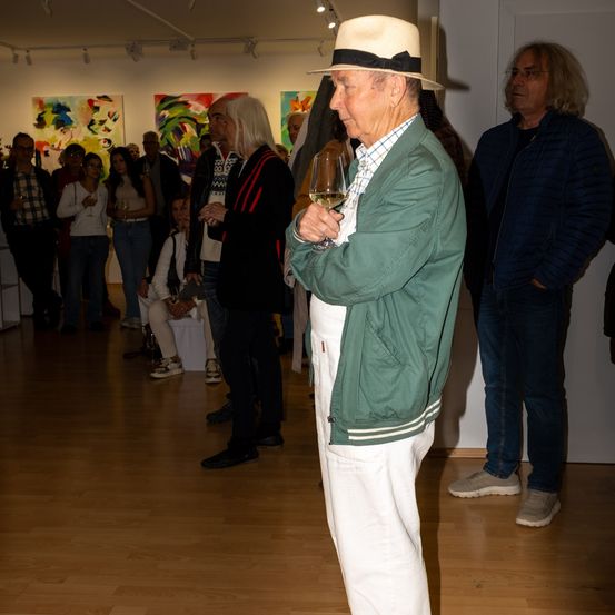 Ein älterer Mann in einem grünen Jackett hält ein Weinglas in einer Kunstgalerie. Er steht neben einem Mann mit Brille und einem anderen mit langen Haaren. Bunte Gemälde schmücken die Wand.