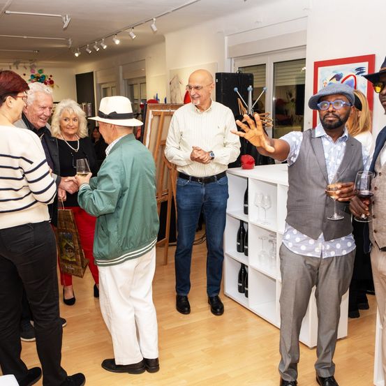 Mehrere Menschen versammeln sich in einer Kunstgalerie. Ein älterer Mann trägt eine grüne Jacke und einen weißen Hut. Ein erwachsener Mann hält ein Weinglas. Flaschen Wein sind auf Regalen ausgestellt.