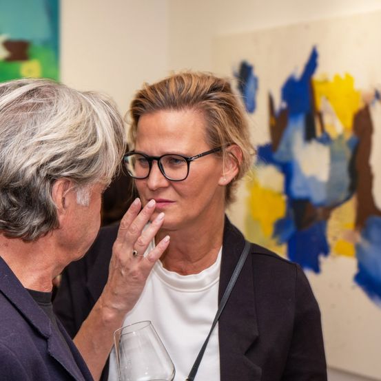 Zwei Frauen in einer Galerie, eine mit Brille, diskutieren über Kunst. Ein Mann hört zu und hält ein Weinglas. Das Gemälde hinter ihnen ist abstrakt mit blauen und gelben Farben.